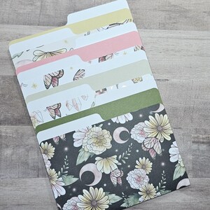 May include: A set of decorative file folders with various patterns. The tabs are white, and the folder designs include butterflies, flowers, crystals, and moons in pastel colors on a dark background. The folders are arranged in a diagonal stack.