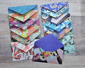 May include: A collection of origami envelopes in various Japanese-inspired patterns. The envelopes feature designs with waves, flowers, and koi fish in shades of blue, orange, purple, and white. The envelopes are arranged on a wooden surface.