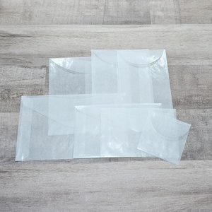 May include: A collection of translucent, white paper envelopes in various sizes. The envelopes are arranged on a light gray wooden surface, with a curved flap closure on each. The paper has a slightly textured appearance.