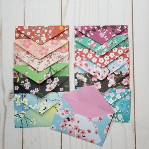 May include: A collection of colorful paper envelopes with floral patterns. The envelopes feature various designs, including cherry blossoms, on a range of backgrounds such as red, pink, green, black, and blue. One envelope is open, revealing a pink interior.