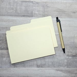 May include: Two cream-coloured file folders with a tab and a pen with a tan barrel and black accents. The pen is lying on a grey wooden surface next to the file folders. The file folders are empty.