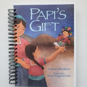 May include: A spiral-bound book titled "Papi's Gift" with a colorful illustration of a girl and a woman. The cover features the text "Papi's Gift" in light blue, with the author's and illustrator's names below.