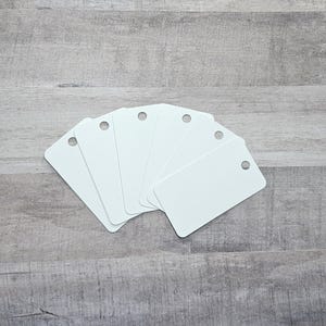 May include: A fan of six blank white rectangular tags with rounded corners and a small hole at the top. The tags are arranged on a gray wooden surface.