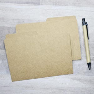 May include: Three tan file folders are stacked on a light wood surface. A matching pen with a black tip and clip rests to the right of the folders. The folders have a simple, clean design.
