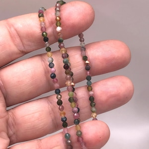 May include: A delicate necklace composed of small, multi-colored gemstone beads. The beads display a variety of colors, including pink, green, yellow, and brown. The necklace is held in a hand, showcasing the intricate details of the jewelry.