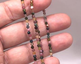 May include: A delicate necklace composed of small, multi-colored gemstone beads. The beads display a variety of colors, including pink, green, yellow, and brown. The necklace is held in a hand, showcasing the intricate details of the jewelry.