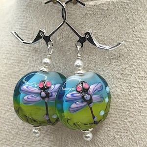 May include: A pair of handcrafted glass earrings featuring a dragonfly design. The round, blue and green glass beads have a dragonfly illustration with pink eyes and lavender wings. Each earring has a silver-colored lever back closure and a pearl bead.