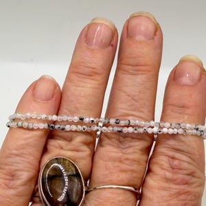 May include: A hand displays a collection of jewelry. Two delicate necklaces of white and black speckled beads are draped across the fingers. A large oval ring with a brown stone and a silver band sits on the ring finger, and a simple silver band ring is also visible.