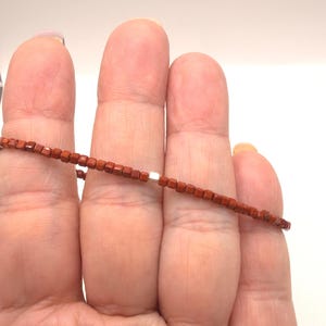 Puede incluir: Un delicado collar con pequeñas cuentas cuadradas de color marrón rojizo ensartadas. El collar tiene un cierre plateado. Las cuentas están espaciadas uniformemente, creando un diseño simple pero elegante. El collar se muestra sobre un fondo neutro.