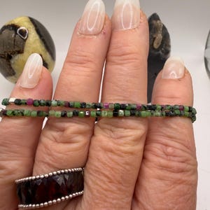 May include: Two delicate beaded bracelets with green, red, and black gemstones. The bracelets are held up by a hand, and a silver ring with a dark red gemstone is also visible. A decorative stone is in the background.