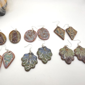 May include: A collection of handmade ceramic earrings in various shapes and designs. The earrings feature a variety of colors, including brown, blue, green, and purple. Some earrings have floral or leaf patterns, while others have geometric shapes. All earrings have silver-colored ear wires.