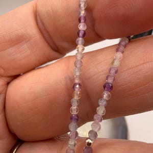 May include: A delicate necklace composed of small, faceted beads in shades of purple, lavender, and clear. The beads are strung together, creating a subtle, elegant accessory. A small gold-colored bead is also visible.