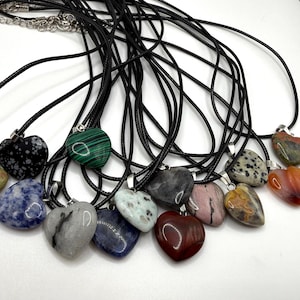 May include: A collection of heart-shaped gemstone pendants on black cords. The pendants vary in color and pattern, including green malachite, speckled gray, and red-brown. Each pendant is attached to a black cord necklace.