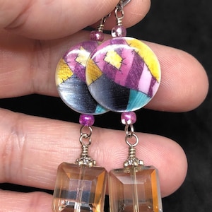 Könnte beinhalten: Ohrringe mit einem bunten Patchwork-Design. Die Ohrringe haben runde Oberteile mit gelben, lila und türkisfarbenen Mustern, die mit rechteckigen, bernsteinfarbenen Kristalltropfen verbunden sind. Kleine lila Perlen und silberfarbene Metallarbeiten vervollständigen das Design.