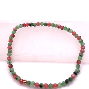 May include: A beaded bracelet featuring small, faceted beads in shades of green, red, and black. The bracelet has a delicate design and a copper-colored accent bead. The beads are strung together to form a flexible band.