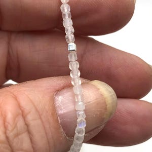 May include: A delicate necklace featuring small, translucent, square-cut beads strung together. A single, small, silver-colored square bead is interspersed within the strand. The beads are held between fingers against a white background.