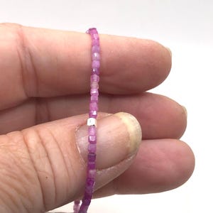 May include: A delicate necklace featuring small, faceted pink gemstones strung together. The beads are a vibrant shade of pink, with a small silver-colored spacer bead. The necklace is held between fingers against a white background.