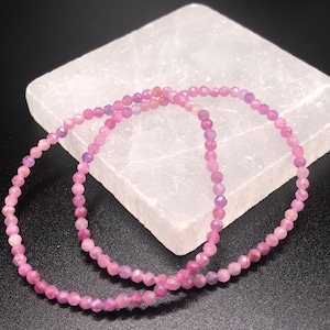 May include: Two pink gemstone bracelets made of small, faceted beads. The bracelets are lying on a white surface.