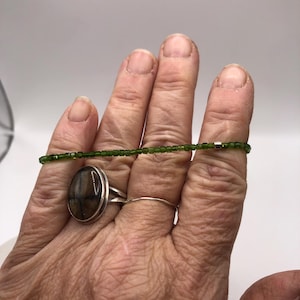 May include: A silver ring with an oval, brown stone and a delicate silver band ring. A green beaded bracelet is stretched across the fingers. The jewelry is displayed against a white background.