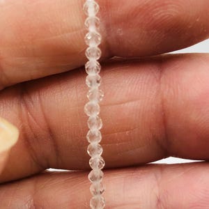 May include: A close-up of a delicate strand of clear, faceted beads. The beads are small and evenly spaced, strung together to create a shimmering, translucent effect. The beads are held between two fingers, highlighting their fine detail and craftsmanship.