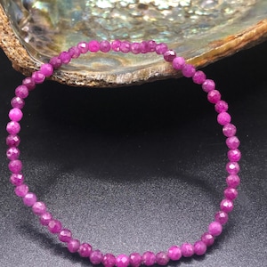 May include: A delicate, magenta-colored beaded bracelet. The bracelet is made of small, faceted beads strung together. The background features a pearlescent, iridescent surface and a dark, solid surface.