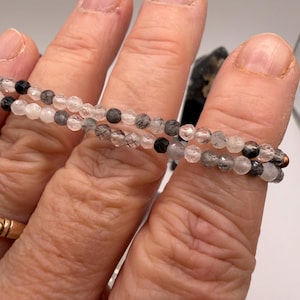 May include: A close-up of two beaded bracelets. The bracelets are made of small, round, clear and black beads. The bracelets are held by a person's fingers. The background is a blurred, neutral color.