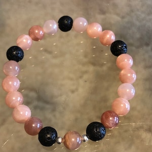 May include: A bracelet made with pink and white sunstone beads and black lava rock beads. The bracelet has silver spacer beads.