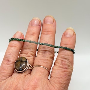 Pode incluir: Uma mão exibe uma pulseira de contas verdes e dois anéis. Um anel é de prata com uma banda fina, e o outro apresenta uma pedra grande, oval, com um padrão em forma de cruz em tons de castanho e preto.