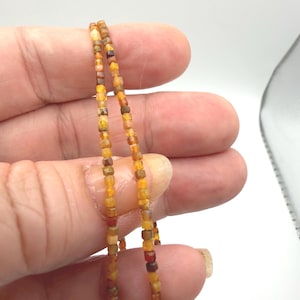 May include: A strand of small, square-shaped beads in varying shades of amber, orange, yellow, and deep brown. The beads are strung together in a continuous line, displaying a translucent quality and a polished, multi-faceted surface texture.