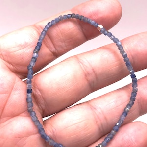 May include: A delicate beaded bracelet featuring small, faceted, square-shaped blue sapphire beads arranged in a continuous loop. A single, small, polished metallic cube bead is integrated into the strand, providing a subtle contrast to the blue.