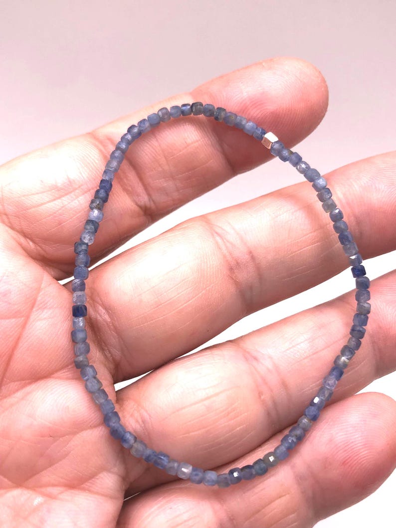 May include: A delicate beaded bracelet featuring small, faceted, square-shaped blue sapphire beads arranged in a continuous loop. A single, small, polished metallic cube bead is integrated into the strand, providing a subtle contrast to the blue.