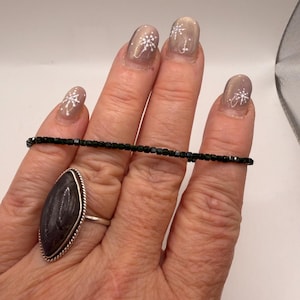 May include: A hand displays a black beaded bracelet and a silver ring with a large, oval, dark gray stone. The ring has a silver rope-like border. The nails are painted with a neutral color and white snowflake designs.