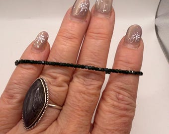 Könnte beinhalten: Eine Hand zeigt ein schwarzes Perlenarmband und einen Silberring mit einem großen, ovalen, dunkelgrauen Stein. Der Ring hat einen silbernen, seilartigen Rand. Die Nägel sind in einer neutralen Farbe mit weißen Schneeflocken-Designs lackiert.