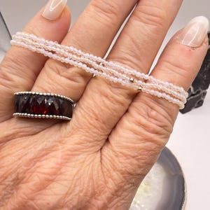 May include: A hand displays a delicate, multi-strand bracelet of small, white beads. A silver ring with a dark red gemstone is on the ring finger. The bracelet is draped across the fingers, showcasing the jewelry.
