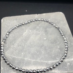 May include: A silver-colored beaded bracelet. The bracelet is made of small, faceted beads strung together to form a circular shape. The beads have a metallic sheen and reflect light. The bracelet is displayed on a white, rectangular surface.