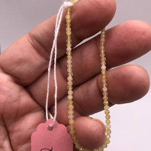 Puede incluir: Una hebra de pequeñas cuentas de color amarillo pálido con un cordón blanco atado. Las cuentas son facetadas y tienen un ligero brillo. El cordón está atado a una etiqueta rosa con las palabras "Gold Opal" escritas en ella.