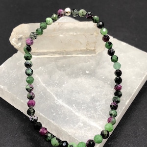 May include: A green, black, and pink gemstone bracelet with a silver bead clasp. The bracelet is made of small, faceted beads and is displayed on a white surface.