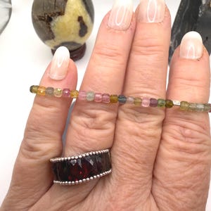 May include: A hand displays a multi-colored tourmaline bead bracelet and a silver ring with a large, faceted, dark red stone. A decorative stone egg and a black crystal are in the background.