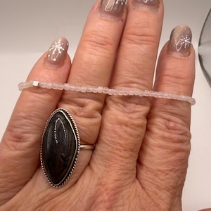 Puede incluir: Un anillo de plata con una piedra oscura ovalada grande y un borde con cuentas. Una pulsera delicada de cuentas transparentes se sostiene en los dedos. Las uñas están pintadas con un esmalte marrón claro y diseños de copos de nieve blancos.
