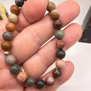 Può includere: Un braccialetto di perline con varie pietre preziose dai toni della terra. Le perline sono nelle tonalità del marrone, beige, grigio e rosa. Il braccialetto è infilato su un cordoncino elastico. Il braccialetto è tenuto in una mano.