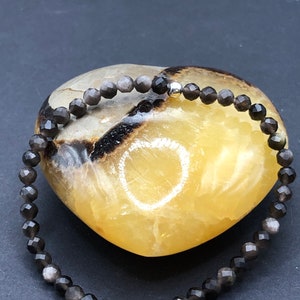 May include: A black gemstone bracelet with a silver bead accent. The bracelet is made of faceted beads and is displayed on a heart-shaped stone.