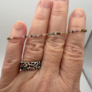 May include: A delicate bracelet composed of tiny, multi-colored tourmaline beads in shades of pink, green, and yellow, and a silver-toned ring featuring an intricate, organic, open-work design with small embedded dark green and clear stones.