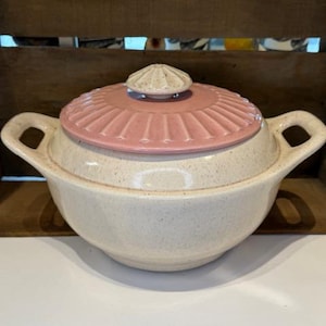 May include: Cream-colored ceramic casserole dish with a pink lid. The lid has a ribbed design and a small, decorative handle. The dish has two handles and a speckled finish, suggesting a vintage or handmade aesthetic.