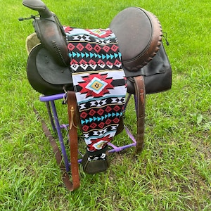 May include: A brown leather saddle with a colorful geometric patterned saddle blanket. The saddle is on a purple metal stand.