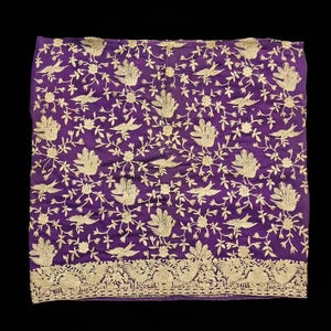 Pode incluir: Um tecido quadrado roxo com um padrão intrincado de pássaros e videiras florais bordadas em fio dourado. A borda inferior apresenta uma faixa horizontal densa de motivos repetitivos de pássaros e folhas, criando um acabamento decorativo.