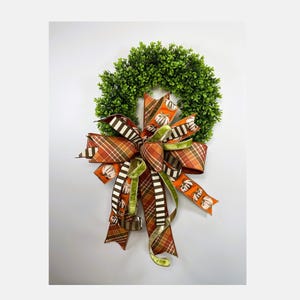 May include: A circular green boxwood wreath adorned with a large decorative bow. The bow features multiple ribbons in orange, brown, and white, displaying patterns of plaid, stripes, and white pumpkin illustrations with long trailing tails.