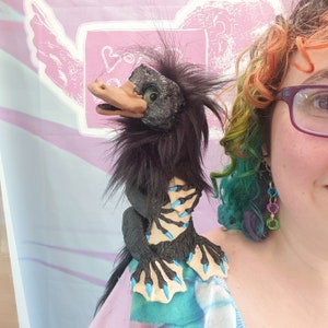 May include: A handcrafted puppet featuring a dark, textured, fur-covered body with a tan, duck-like bill and large, expressive eyes. The creature has webbed, black hands and feet with bright blue claws, and is designed to be worn on the shoulder.