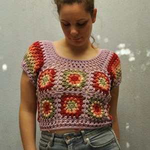 May include: A woman wearing a purple crochet top with a floral pattern. The top has short sleeves and a square neckline. The woman is wearing blue jeans.