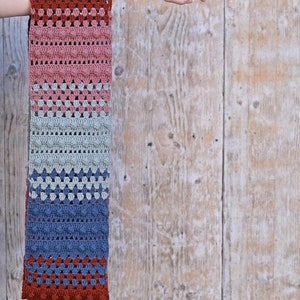 May include: A crocheted scarf with a multi-colored striped pattern in shades of red, pink, green, blue, and gray. The scarf has a textured design with small squares and bobbles.
