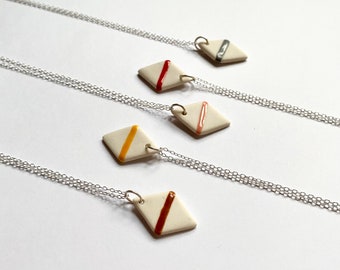 May include: Four necklaces with white ceramic diamond-shaped pendants featuring a single colored line. The lines are red, yellow, pink, and gray.